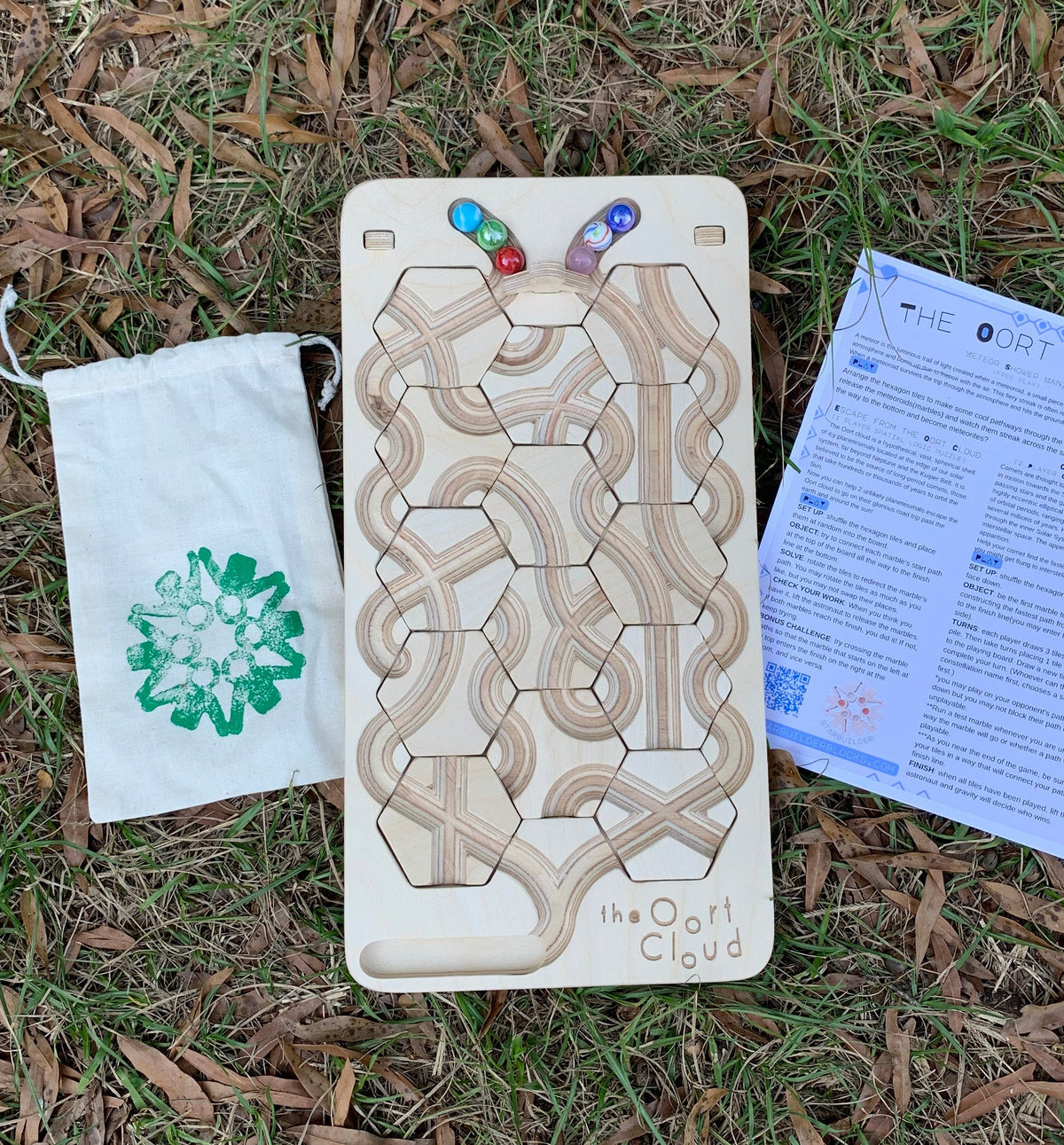 The Oort Cloud – Wooden Marble Run Puzzle, featuring a handmade board with marble tracks and hexagonal tiles, a small cloth bag of marbles, and an instruction sheet, all set on a grass lawn.