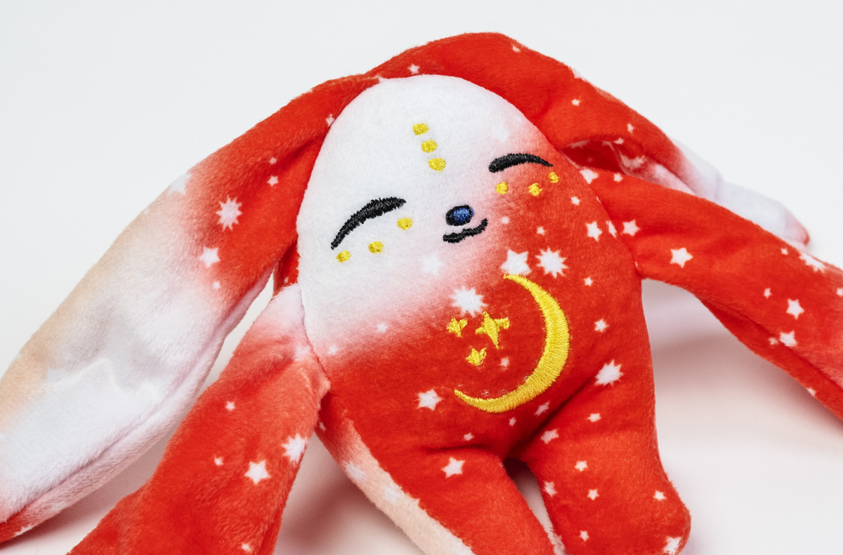 The Rover Mini Wrist Hugger – Magnetic Sensory Plush is a red, pocket-sized toy with long ears, a white face, closed eyes, and a yellow embroidered crescent moon and stars pattern designed as a magnetic wrist accessory.