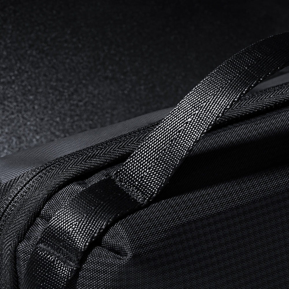 Close-up of the black Tech Pouch with textured fabric and a strong woven strap, displayed on a dark, reflective surface. The image showcases detailed stitching and high-quality materials, emphasizing its role as a travel organizer.