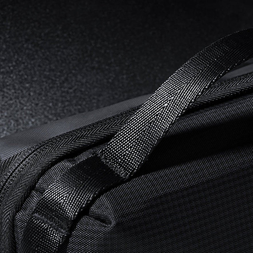 Close-up of the black Tech Pouch with textured fabric and a strong woven strap, displayed on a dark, reflective surface. The image showcases detailed stitching and high-quality materials, emphasizing its role as a travel organizer.
