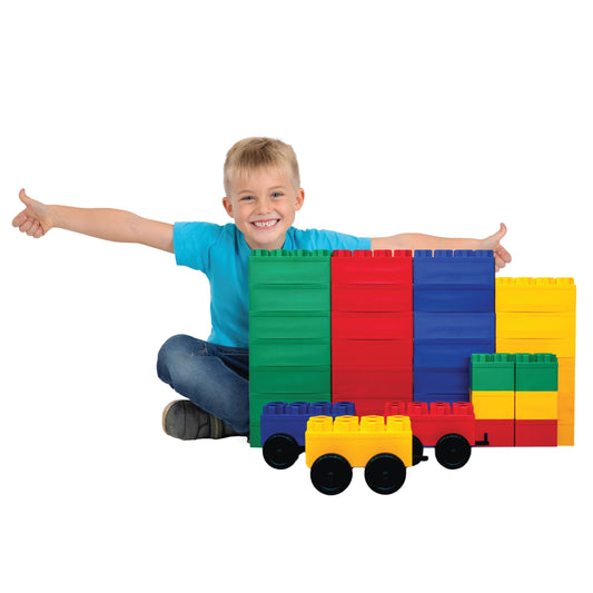 A smiling child in a blue shirt sits on the floor, arms outstretched behind a set of Jumbo Building Blocks 36 PC with 3 Wheel Kits - Train Set, including colorful giant blocks and a toy car built from the same pieces.