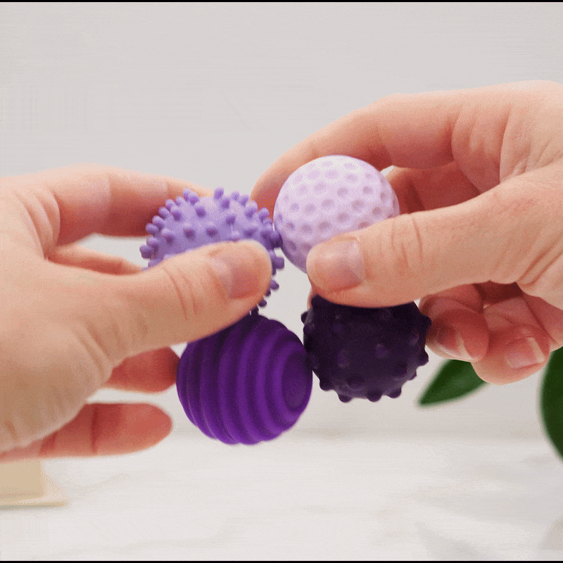 Two hands hold four Signets sensory balls in shades of purple, each featuring unique textures with raised bumps and ridges for varied fidgeting. A blurred green plant in the background adds a calming touch to enhance focus.
