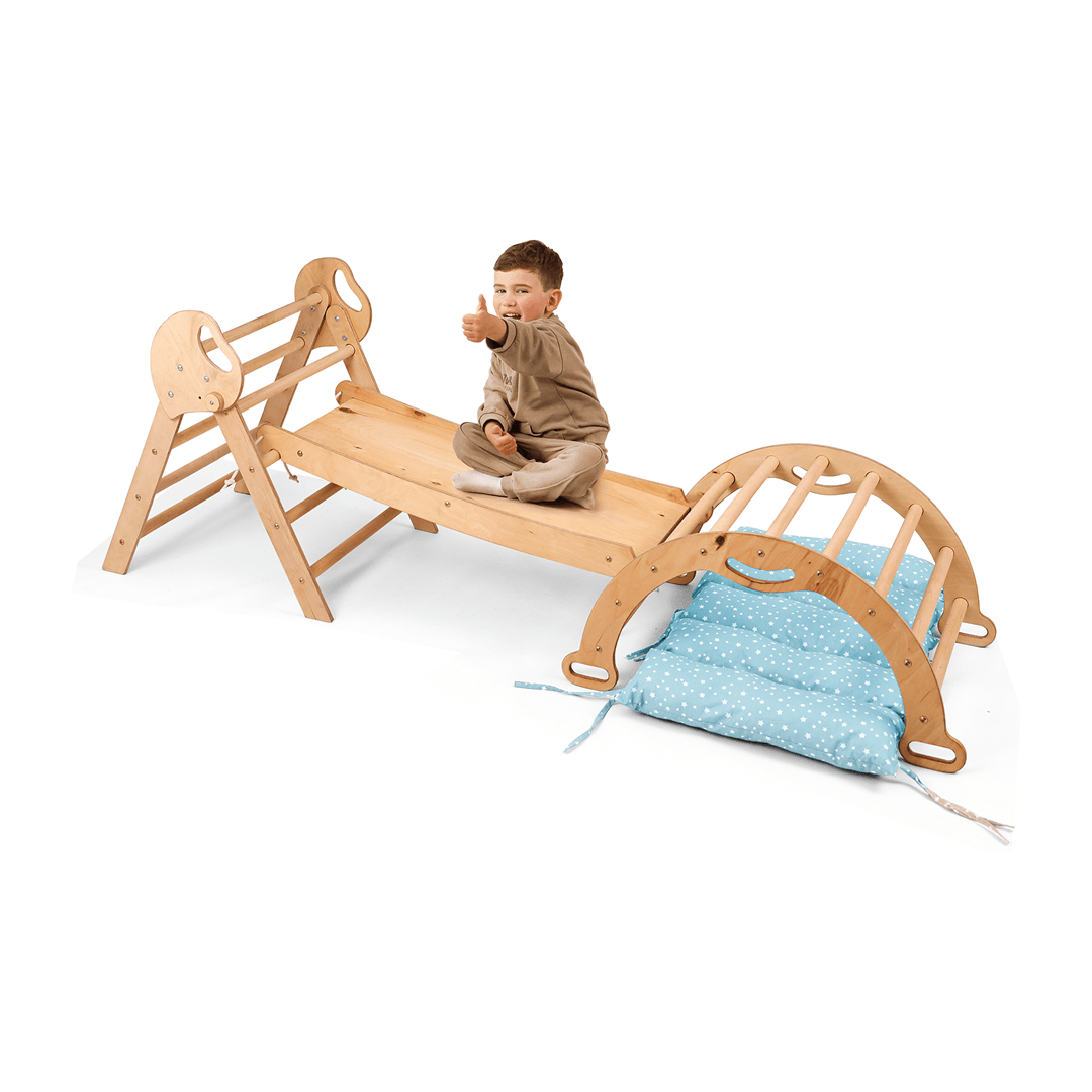 A toddler sits cross-legged on the beige 4in1 Montessori Climbing Set—Pikler Triangle, Arch, Slide Board, and Cushion for ages 1–3—with a thumbs-up. The arch and blue cushion are nearby, all set against a white background.