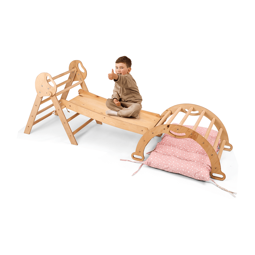 A young child sits cross-legged on a wooden play structure, giving a thumbs-up next to the beige 4in1 Montessori Climbing Set: Pikler Triangle, Arch, Slide Board, and Cushion for Toddlers (1–3 Years); pink polka-dot cushions complete the setup. The background is white.