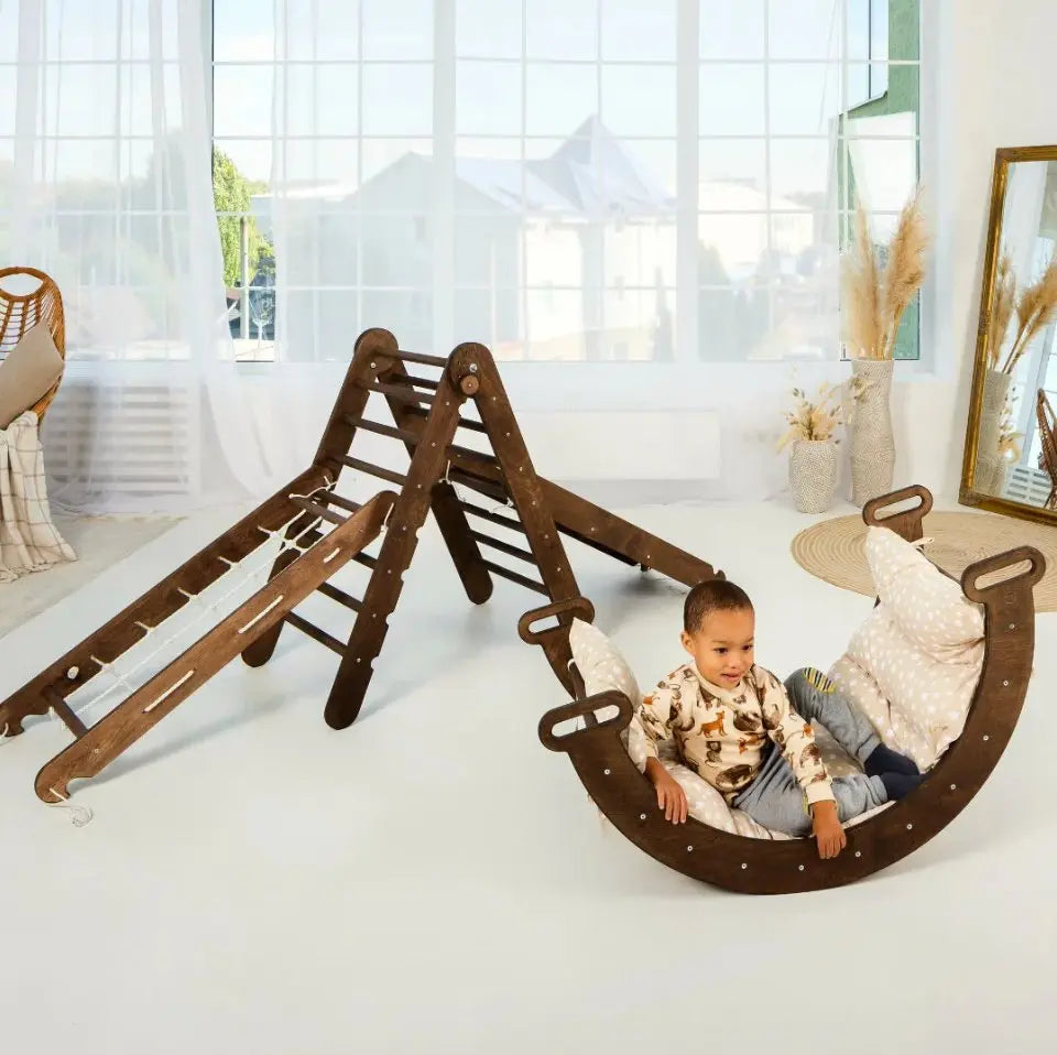 A young child sits at the base of a Chocolate 5in1 Montessori Climbing Set—featuring a Triangle Ladder, Arch/Rocker, Slide Board/Ramp, Netting Rope, and Cushion—surrounded by light pillows in a bright, cozy room with large windows.