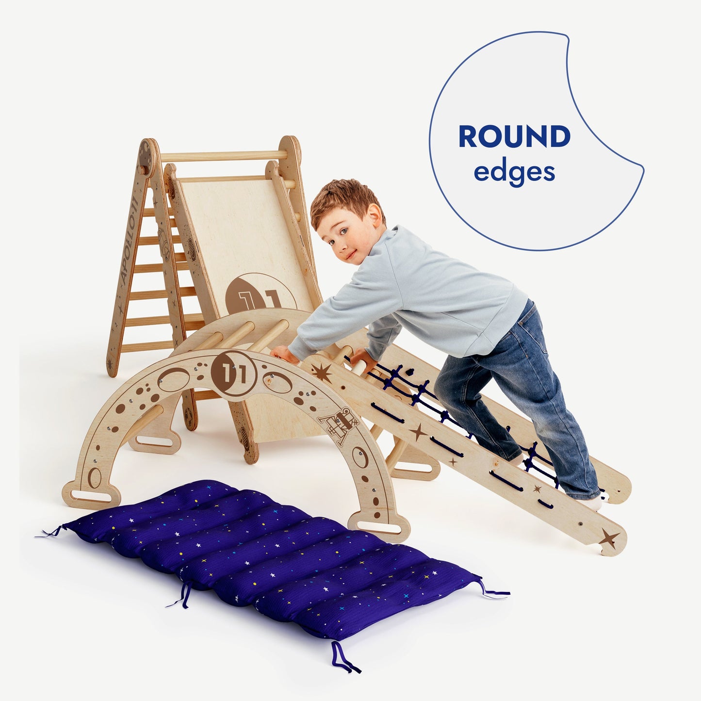 A young boy climbs the Apollo 5in1 Climbing Set: Pikler Triangle, Net, Arch, Slide Board, and Cushion for Little Astronauts. The image shows the blue padded mat on the floor and a ROUND edges label. The set is made of light wood.