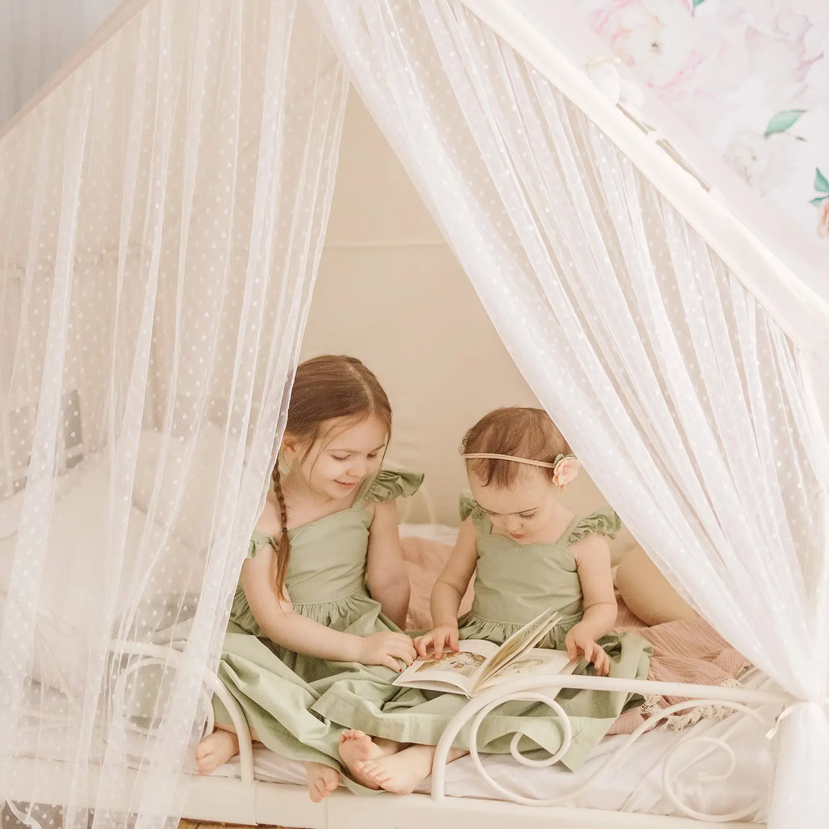 Two girls in matching green dresses sit inside a Large Cozy Cabin Play Tent with Star Lights, a white, sheer-curtained reading nook, and read a book together.