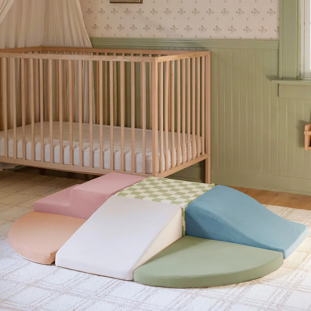 PlayNest Soft Climbing Blocks & Ball Pit Set – Morandi consisting of pastel-colored foam blocks, including a ramp, stairs, and curved floor mats, arranged on a rug to create an indoor playground in front of a crib.