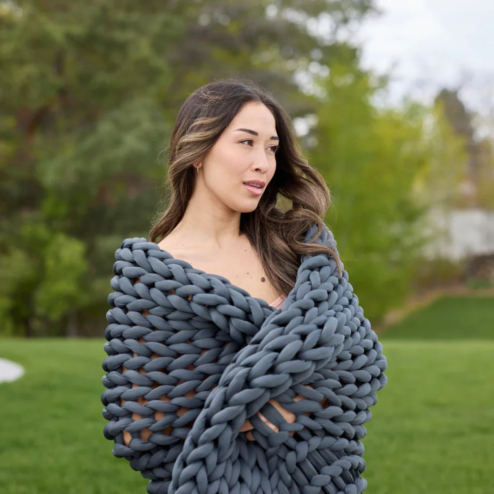 A woman with long brown hair stands outdoors, wrapped in the thick, dark gray Ice Knit Weighted Blanket, surrounded by green grass and trees.
