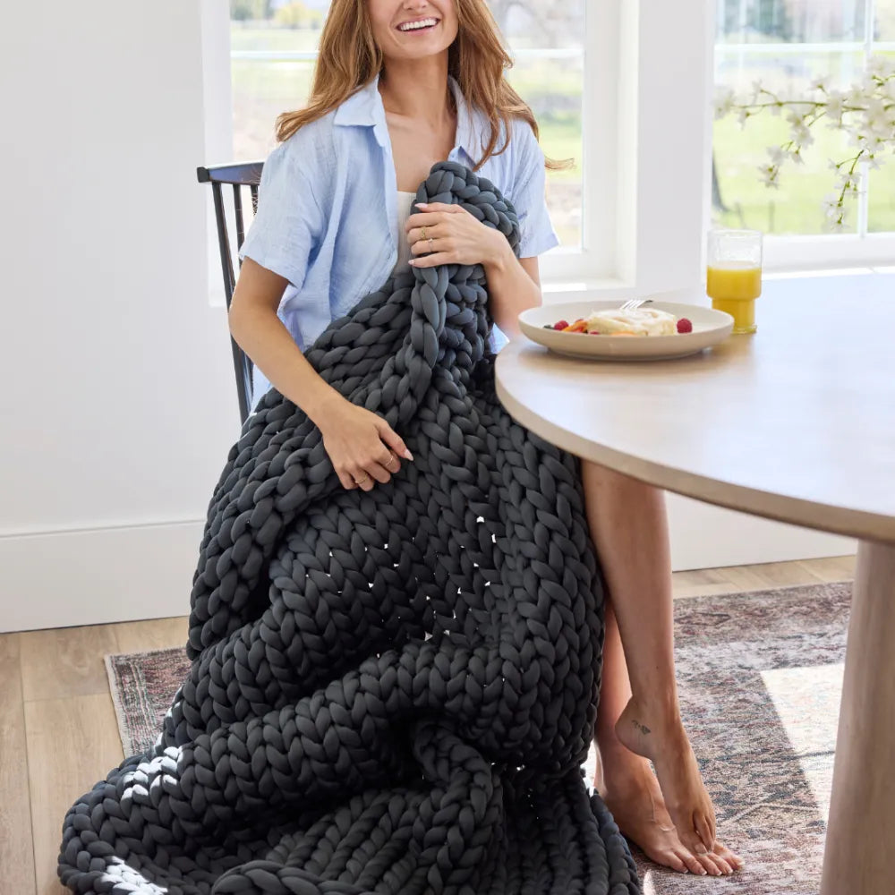 A woman smiles at a dining table with food and orange juice, wrapped in a chunky dark gray Ice Knit Weighted Blanket as sunlight streams through large windows in the background.