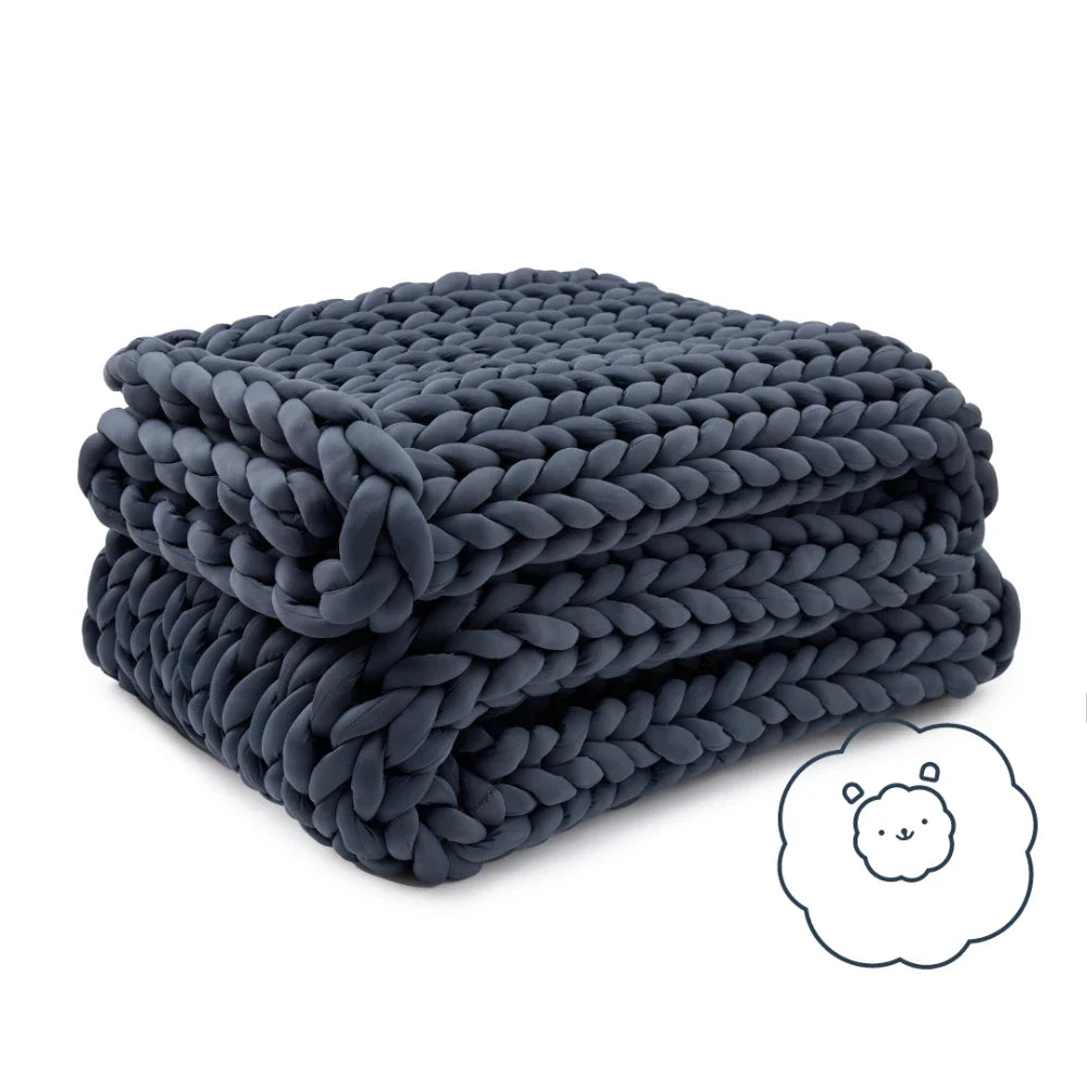 A chunky, dark gray Ice Knit Weighted Blanket is folded neatly. In the bottom right corner, a small illustration of a smiling sheep in a fluffy cloud hints at this breathable blanket’s cozy comfort.