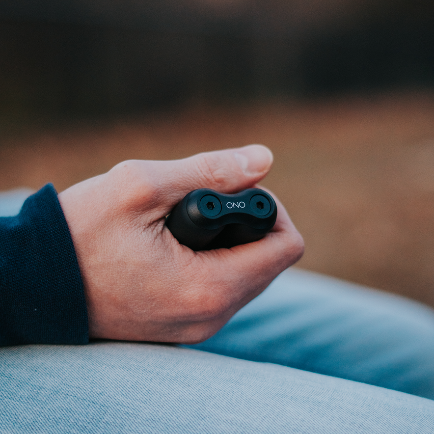 A hand holds a small black ONO Roller Soft Touch Plastic, thumb on top and fingers wrapped around it. This silent fidget toy is ideal for stress relief, shown against a blurred outdoor background.