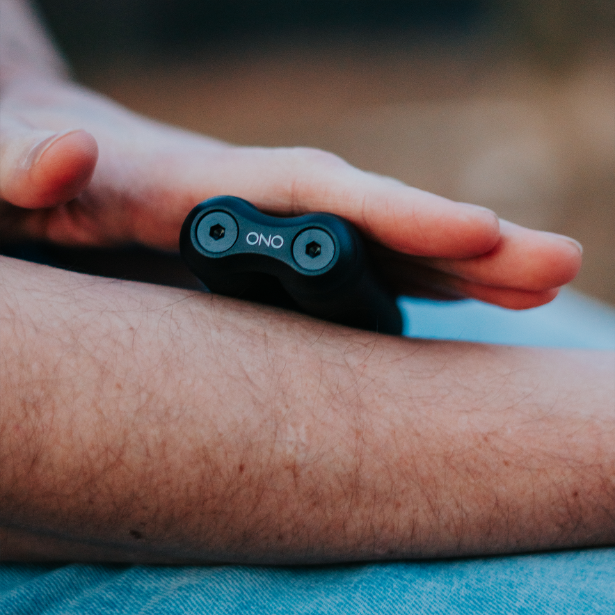 A close-up of a person’s hand holding the ONO Roller Soft Touch Plastic—a small black device with two visible screws and a smooth, rounded surface—against their bare forearm for stress relief.