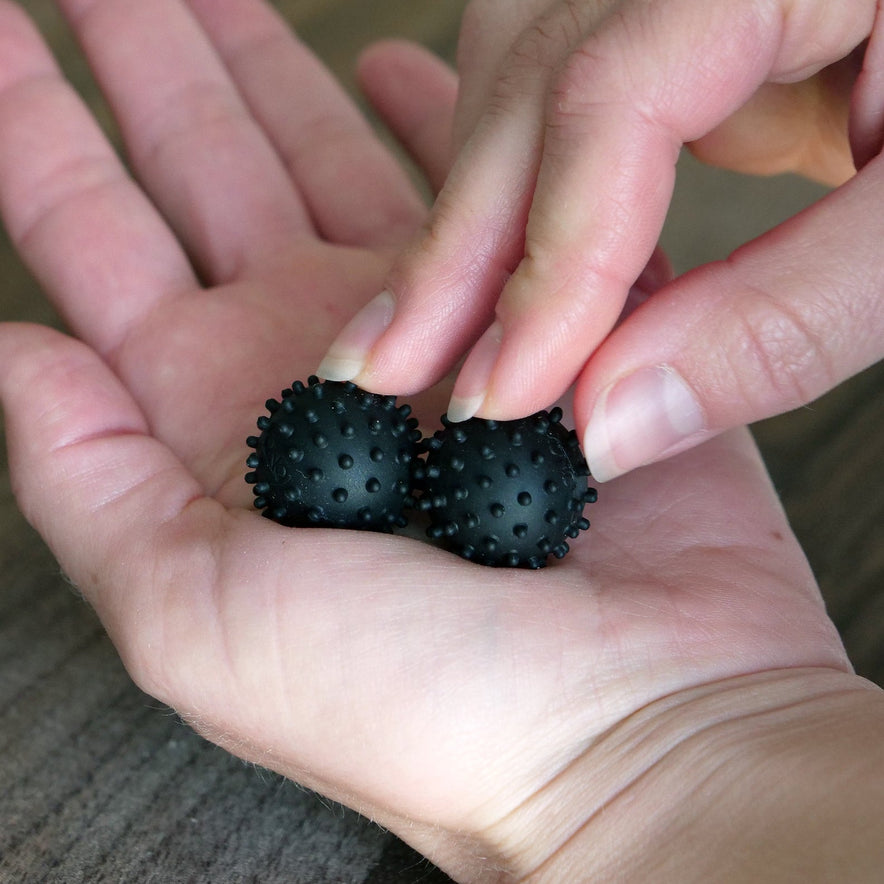 A hand holds two ONO Sigbits, small black spiky massage balls popular as fidget toys, while another hand reaches to pick one up. The background is a wooden surface.
