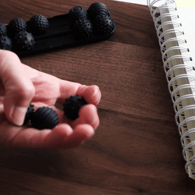 A hand holds two ONO Sigbits—small, textured black fidget balls ideal for sensory stimulation or focus—over a wooden desk. More ONO Sigbits rest in a nearby tray next to a spiral notebook.