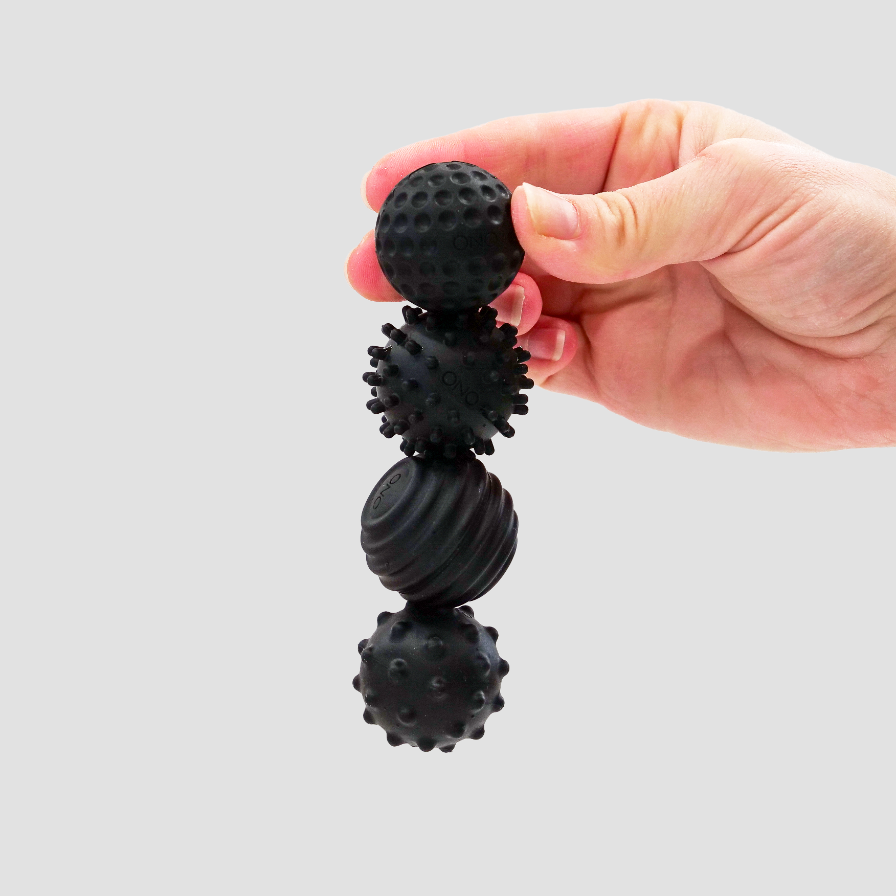 A hand holds four black Signets massage balls, each with a unique surface texture, stacked vertically against a plain light gray background, providing an engaging fidgeting experience.