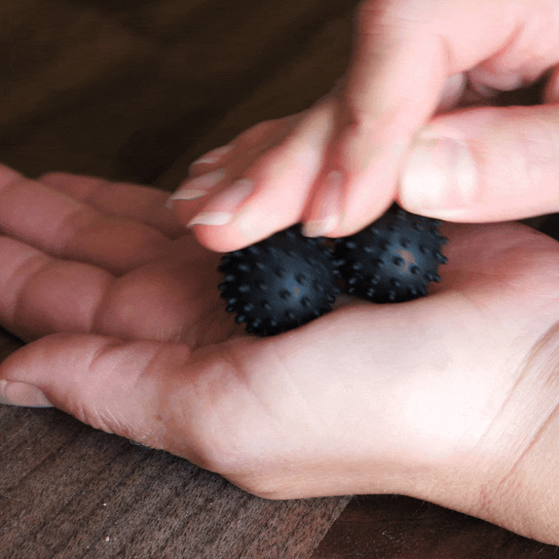 A person rolls two ONO Sigbits between their palms on a wooden surface, demonstrating a hand massage technique that also serves as a sensory stimulation tool and focus enhancer.