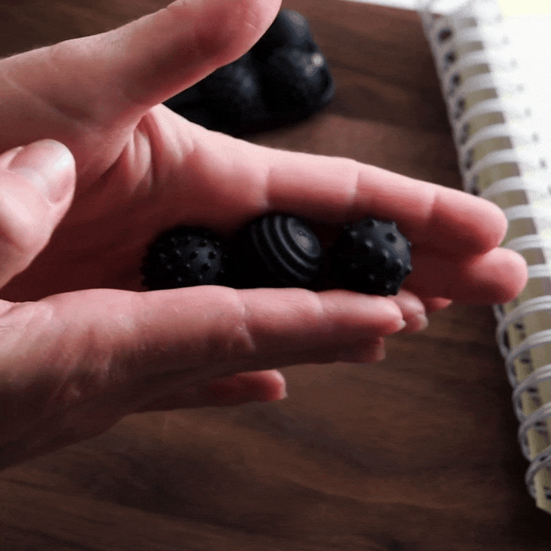 A hand rolls three unique black ONO Sigbits fidget beads over the fingers, providing sensory stimulation and discreet focus enhancement against a blurred background with a notebook and wooden surface.