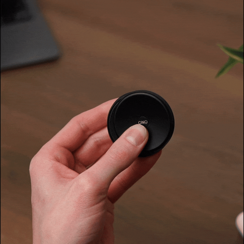 A hand presses the center of the ONO Slider Aluminum, a small, round calming fidget toy, making its outer ring glow blue. A laptop and a plant are visible blurred in the background.