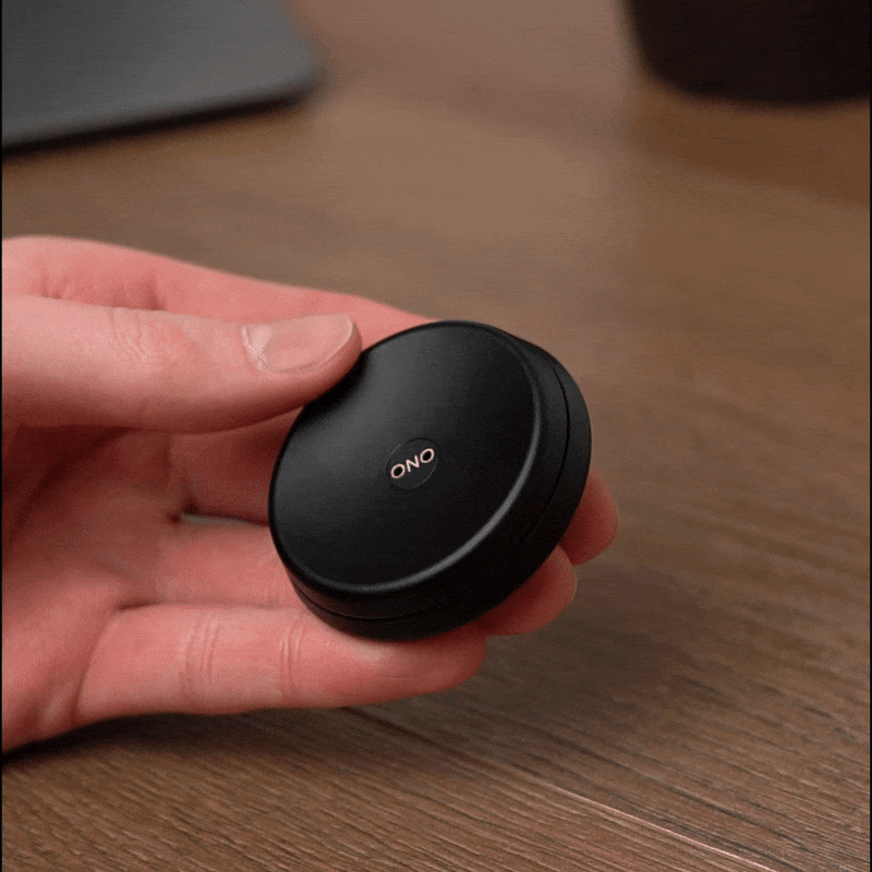 A hand holds a small, round, black ONO Slider Aluminum device labeled ONO. Twisted open, it reveals a calming fidget toy case containing white wireless earbuds, resting on a wooden surface.