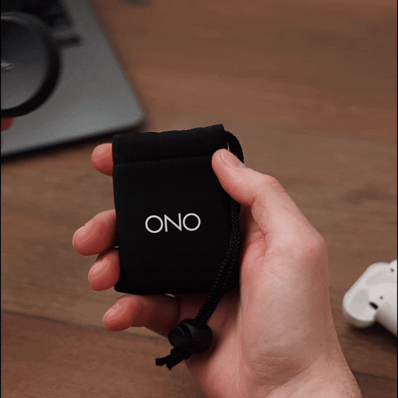 A hand holds a small black drawstring pouch labeled ONO in white letters, with a wooden surface, part of a laptop, and wireless earbuds in the background—ideal for carrying your ONO Slider Aluminum calming fidget toy.