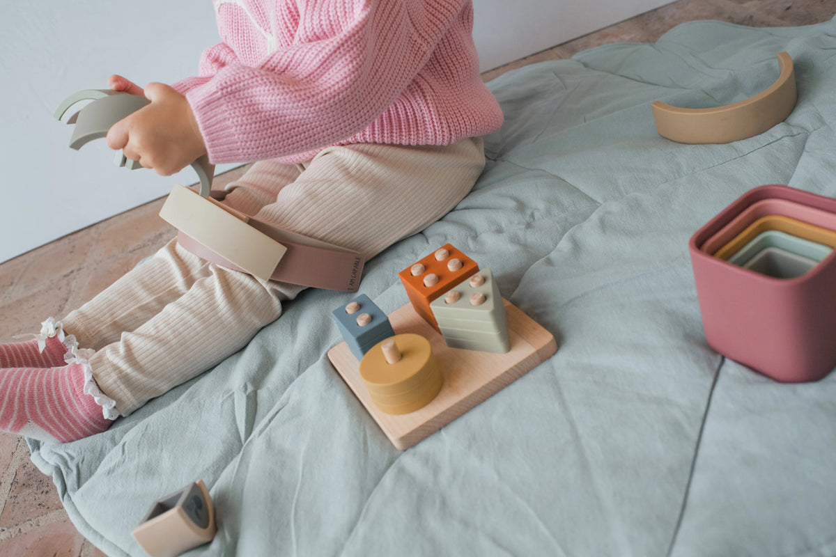 A child sits on a mat engaging in sensory play with a Montessori First Birthday Gift Set – 3 Toys, featuring a stacking toy, geometric shapes on a wooden base, and a set of nesting cups.