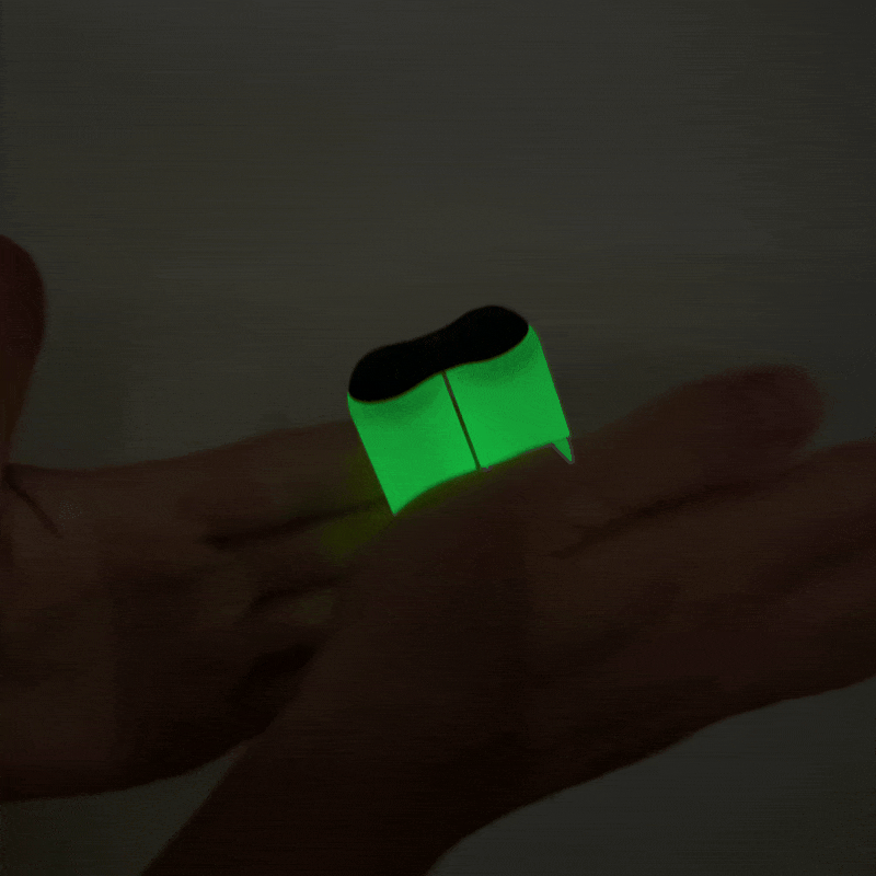 Two hands hold an ONO Roller Soft Touch Plastic, glowing green with a black top, in a dark setting. The toy emits bright light, illuminating the hands and surroundings—ideal for stress relief.