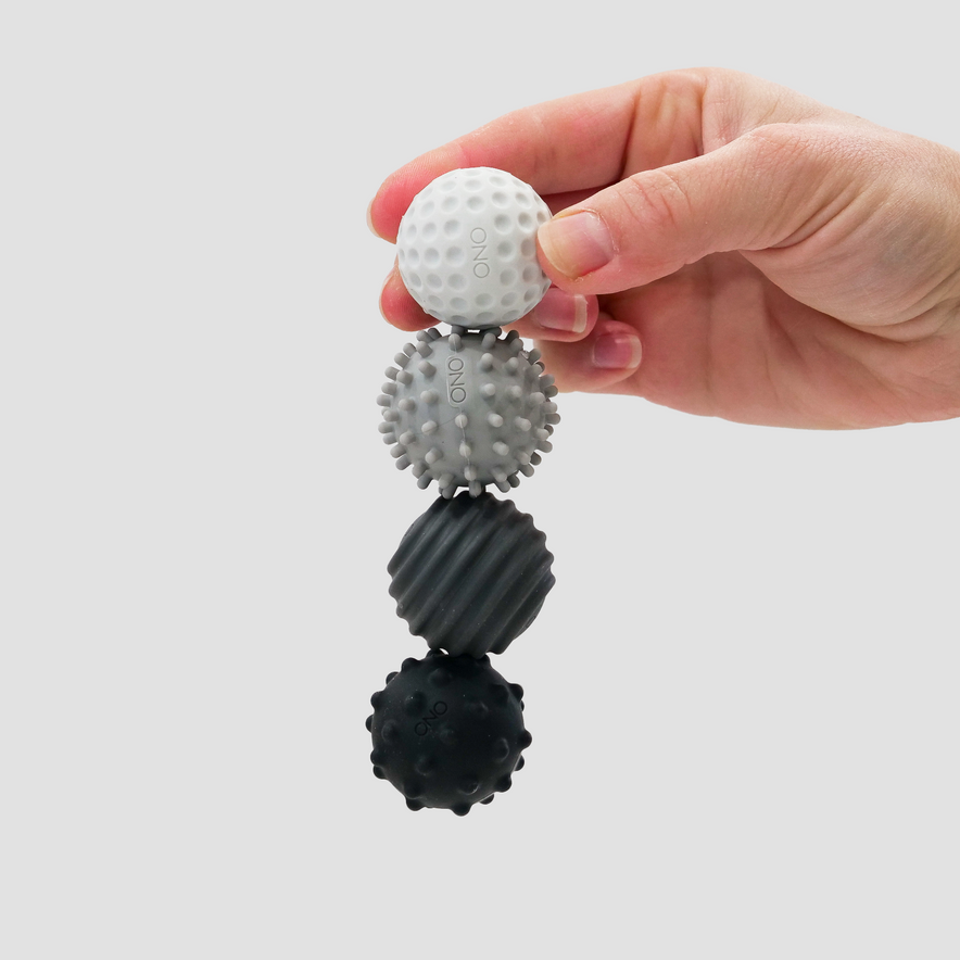 A hand holds four Signets—silicone-coated magnetic balls, each a different shade of grey—stacked vertically against a plain background. These stylish fidget tools offer a unique tactile experience to enhance focus.