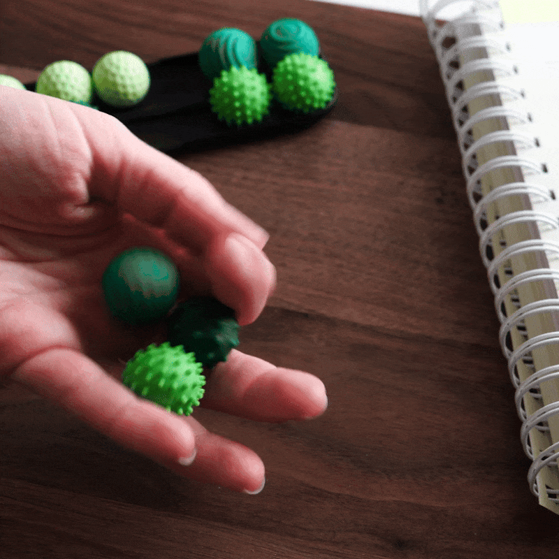 A hand holds small green ONO Sigbits stress balls—ideal fidget toys—over a wooden desk with a spiral notebook and more ONO Sigbits in the background.