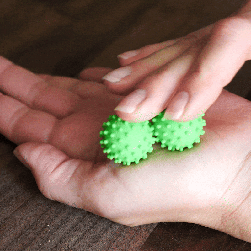 A hand rolls two ONO Sigbits—small, spiky green massage balls—across another palm on a wooden surface, providing calming sensory stimulation and serving as a discreet fidget toy.
