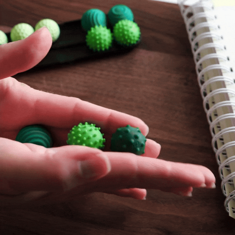 A hand holds two ONO Sigbits—small textured green sensory balls, one smooth and one spiky—over a wooden surface. These fidget toys sit near similar balls and a spiral notebook in the background.