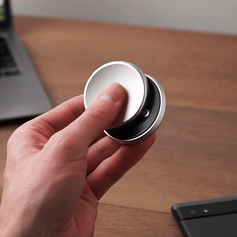 A hand spins the ONO Slider Aluminum, a round silver calming fidget toy with magnetized disks, on a wooden surface near a laptop and smartphone.
