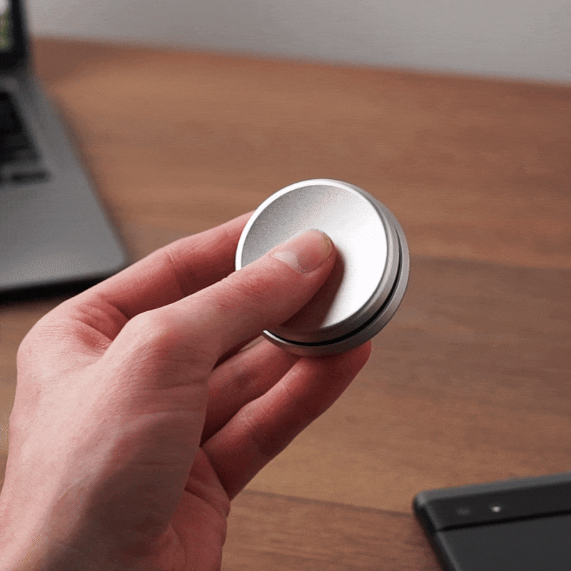A hand spins the ONO Slider Aluminum, a calming magnetic fidget toy, on a wooden desk beside an open laptop and a smartphone.
