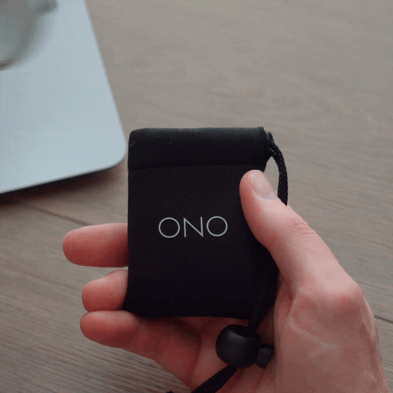 A hand holds a black pouch labeled ONO, then pulls out a compact ONO Slider Aluminum foldable magnifying glass and opens it to reveal its circular lens, with a wooden surface and part of a laptop in the background.