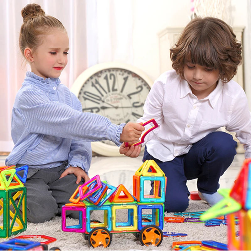 On the floor, two children engage with the Giant Magnetic Building Blocks Set for Kids, a colorful STEM learning tool. A large clock is visible behind them. The girl in a blue blouse hands a block to the boy in a white shirt; both are smiling while playing with these safe and durable toys.
