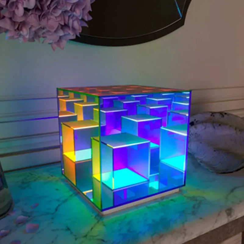 The Vibrant RGB LED Night Light for Parties and Home Décor casts a dynamic, geometric, translucent cube design with vibrant blue, green, yellow, and purple colors on a marble surface. The 3D maze-like reflections create striking visuals against backdrop LED decoration and flowers.
