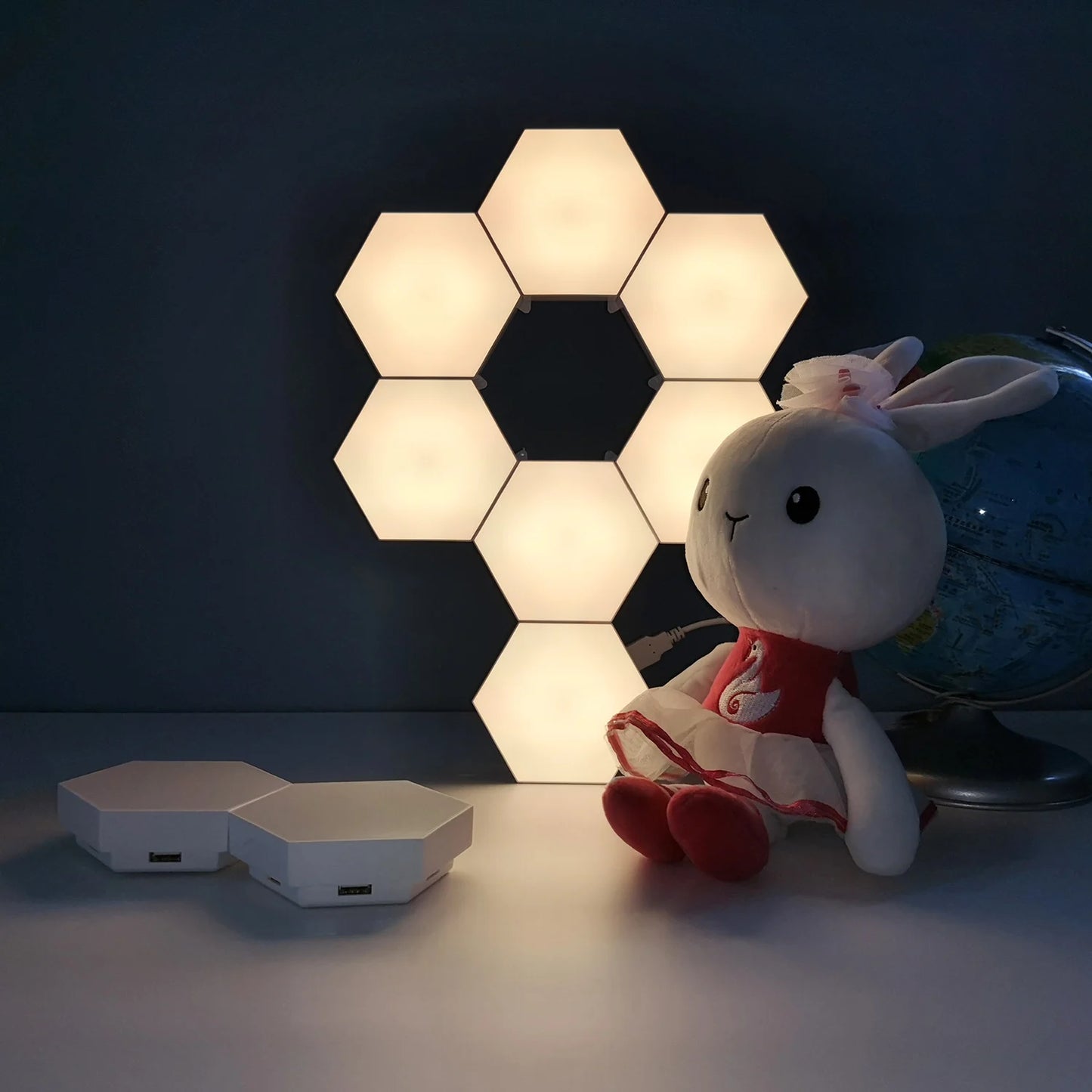 Modular Hexagonal LED Wall Panels provide a calming glow on a dark wall. Nearby, a plush rabbit toy in a red dress is placed beside a globe on the shelf, and a small color-changing hexagonal device with remote capabilities complements the scene in the foreground.