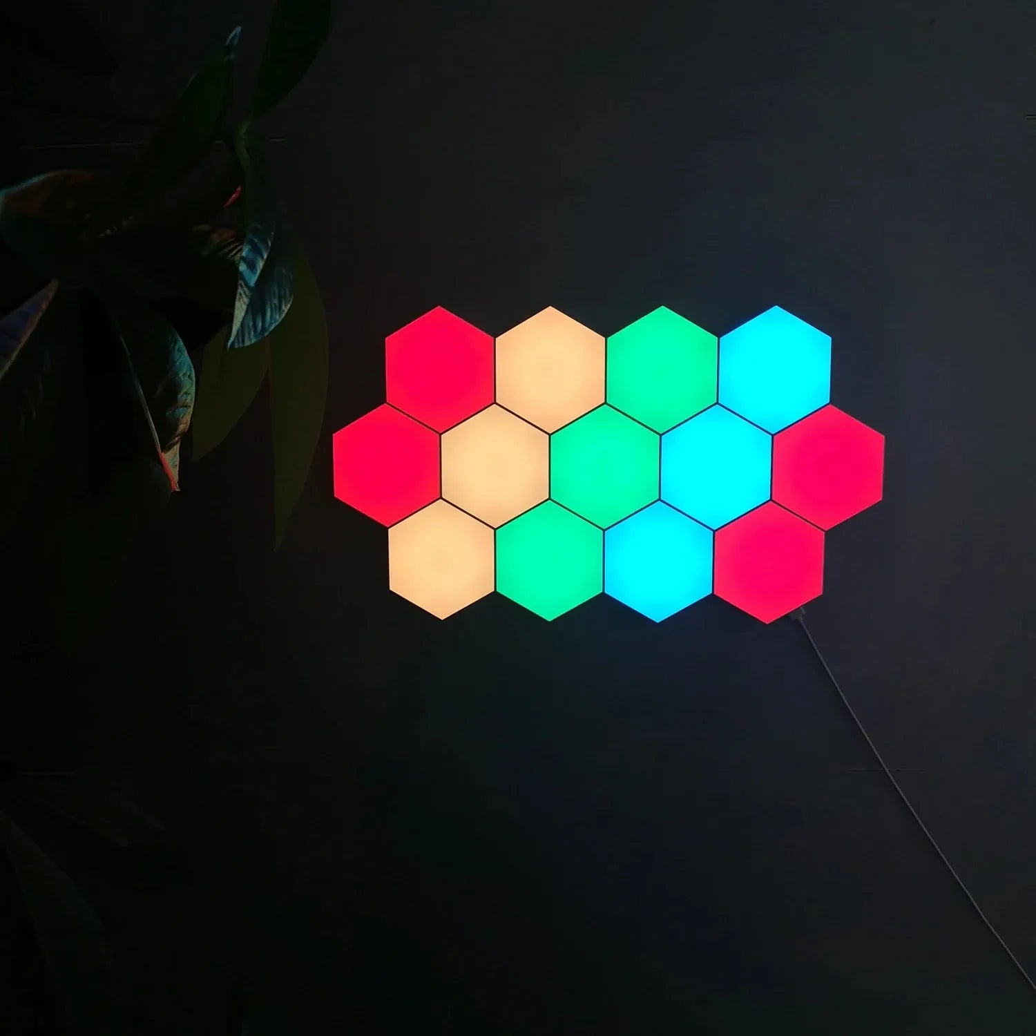 Twelve Hexagonal LED Modular Night Light Panels for Sensory Rooms, glowing in red, yellow, and blue-green, are mounted on a dark wall with a cable at the bottom right and leaves partly visible on the left.