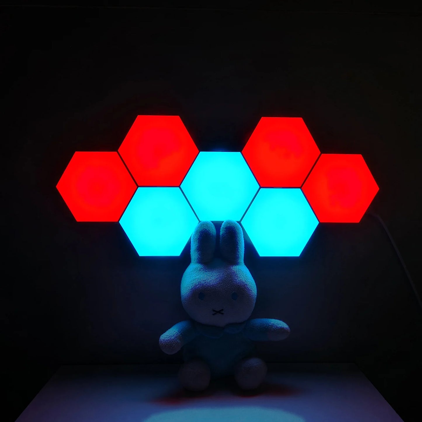 A plush rabbit is placed in front of Modular Hexagonal LED Wall Panels, which have color-changing tiles illuminated in alternating red and blue, creating a vibrant backdrop against the dark background, perfect for mood-setting as a night light.