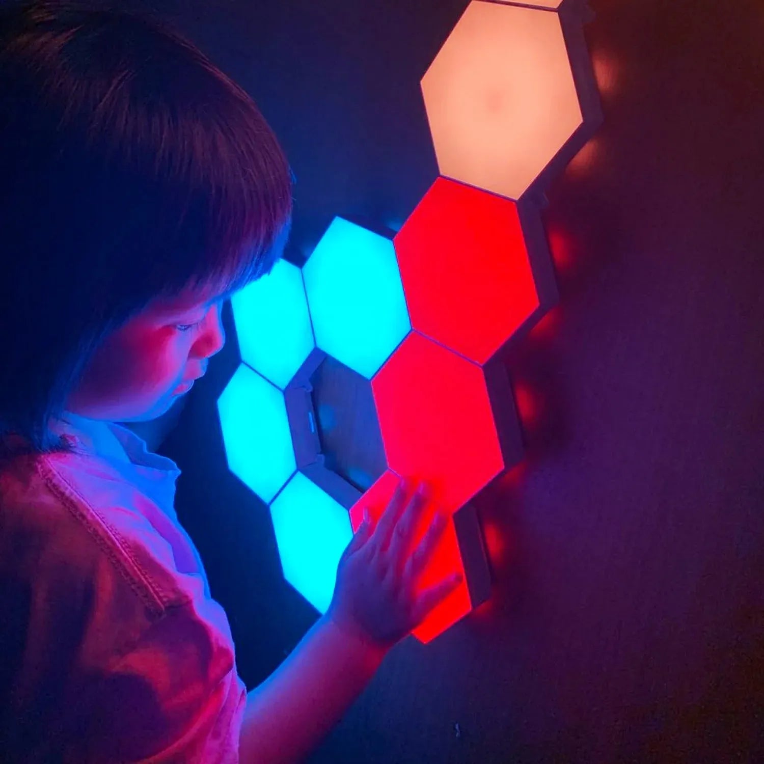 A young child touches a Hexagonal LED Modular Night Light Panel for Sensory Rooms, watching calming blue, red, and white lights cast colorful glows on their face in the dark—a soothing lamp ideal for autism support.