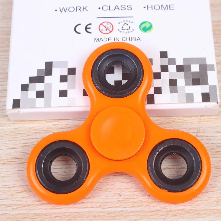 The Premium ABS Tri-Spinner Fidget Toy for stress relief, ideal for autism and ADHD, is colored orange with three black circles. It sits on a box labeled Work Class Home and Made in China, featuring recycling symbols against a wooden surface background.