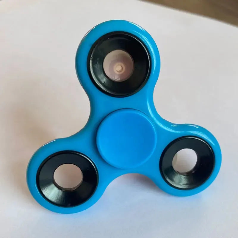 A light blue Premium ABS Tri-Spinner Fidget Toy with three rounded arms and black metallic circles rests on a white surface, providing stress relief through its soothing motion. Ideal for all ages, its perfect for Autism and ADHD.
