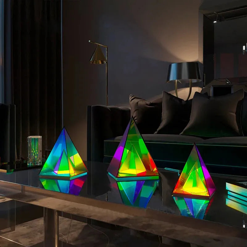 Three vibrant RGB LED pyramid lights, part of the Vibrant RGB LED Night Light collection, sit on a reflective glass table, emitting hues of green, blue, yellow, and orange. Behind them are a dark sofa, pillows, lamp, and curtains that enhance the cozy atmosphere.