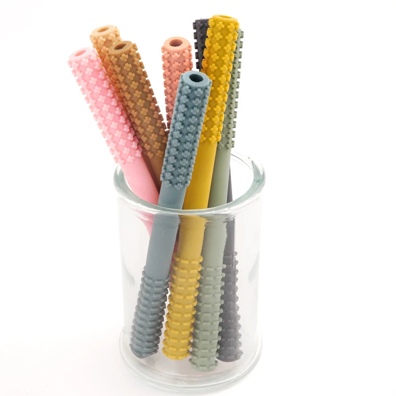 A glass jar contains seven textured silicone straws in pink, brown, yellow, teal, and gray. Designed with a unique bumpy grip for small hands, these straws double as the Essential Tiny Teething Toy for Infants, providing safe chewing fun while protecting the oral cavity.