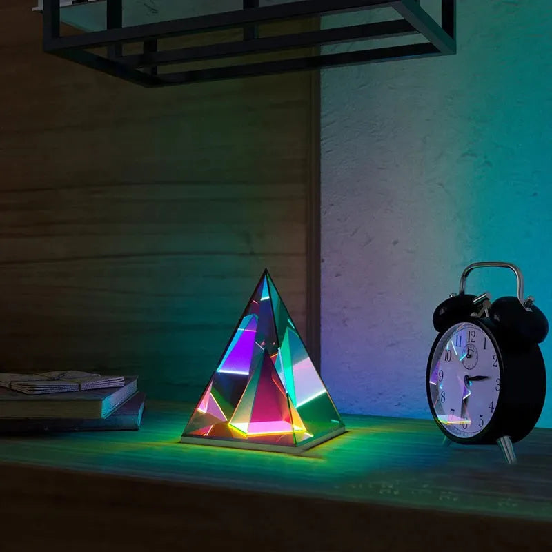 A Vibrant RGB LED Night Light casts colorful light on a wooden table, accompanied by a classic black alarm clock. The dimly lit setting features partially visible books in the background.
