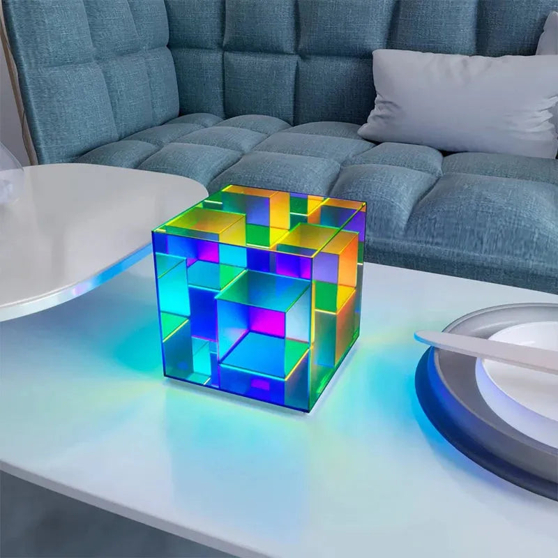 A Vibrant RGB LED Night Light emits a soft glow on a white table, its surfaces reflecting a spectrum of colors. Behind it is a blue sofa with cushions, while the table also holds a white circular object and a plate with a utensil.