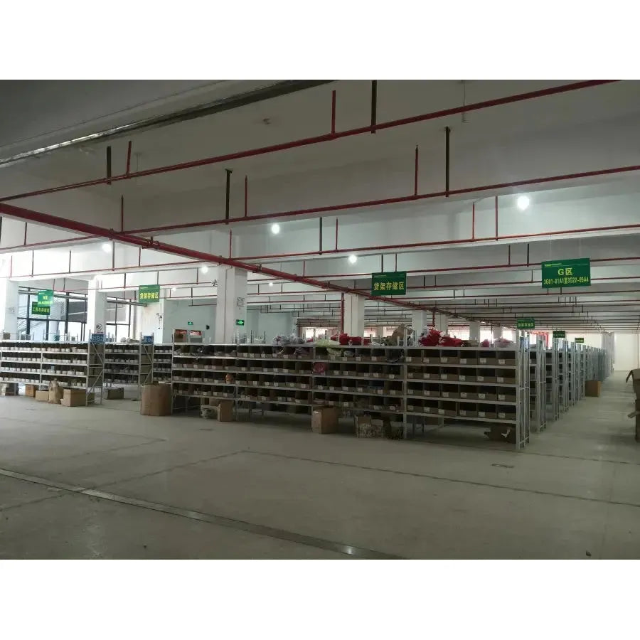 A warehouse interior features rows of metal shelves stocked with items like the Koala Night Light Plush Toy, a musical sensory sleep aid for newborns. Green signs and exposed pipes and lights decorate the ceiling, while the clean floor remains empty.
