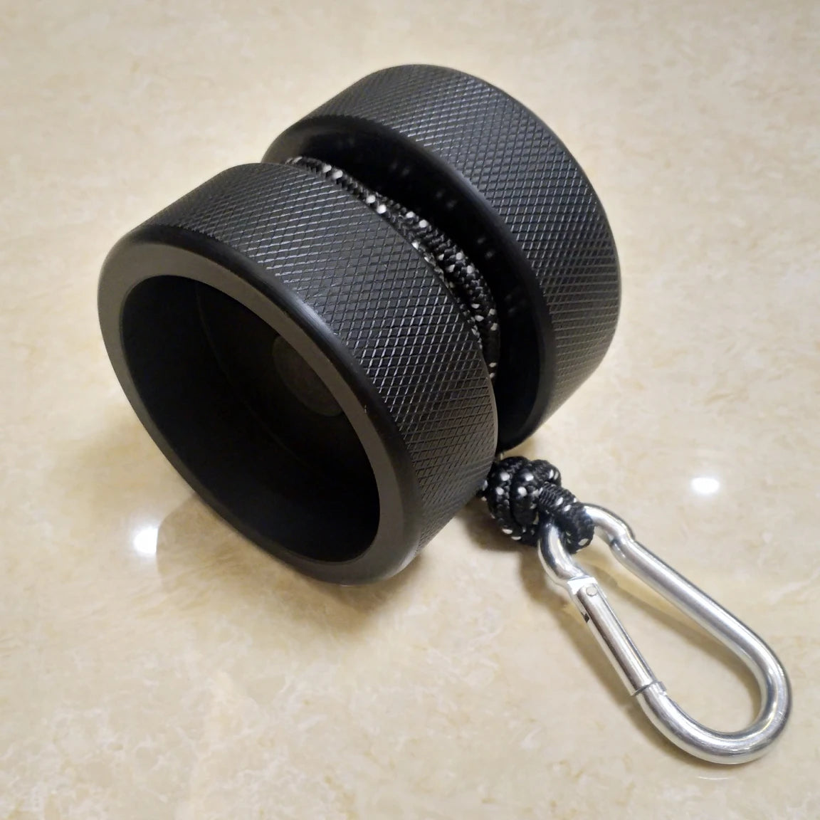The Finger Grip and Forearm Strength Trainer is a black metal ring with a textured surface, attached to a rope and silver carabiner for an anti-slip grip, enhancing finger control. It rests elegantly on marble, showcasing its robust and functional design.