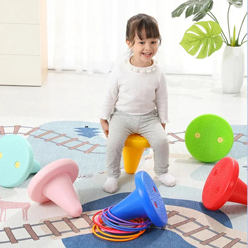 A young child sits on a small, colorful chair similar to the Kids Balance Training Unicorner Chair with Throwing Ring in a playroom. The room has a rug with train tracks and various colorful stools and rings for balance training. A large leafy plant stands near the window in the background.