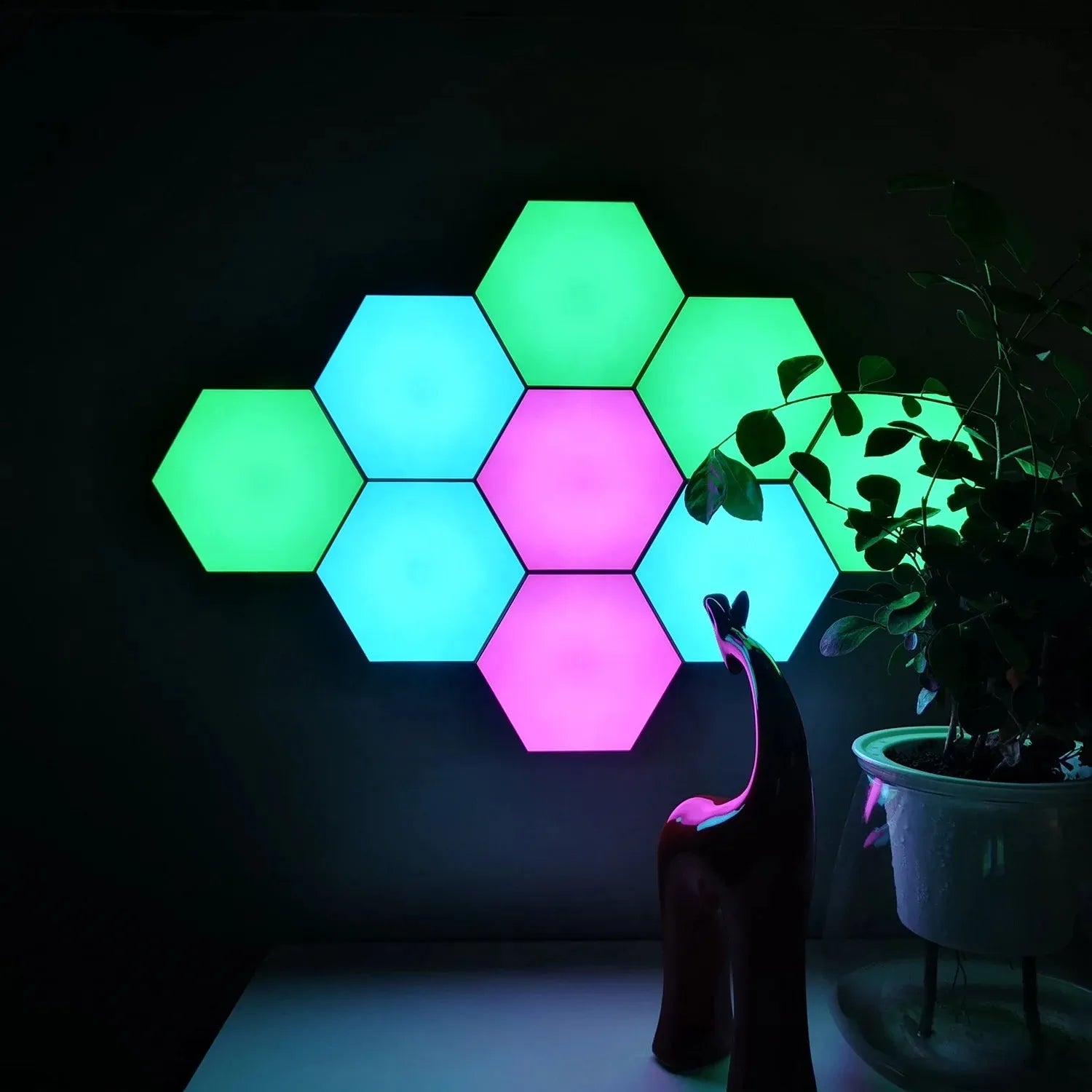 Hexagonal LED Modular Night Light Panels for Sensory Rooms glow in green, blue, and pink on a dark wall, offering a stylish color-changing night light. A leafy potted plant and a modern red figurine decorate the nearby surface.